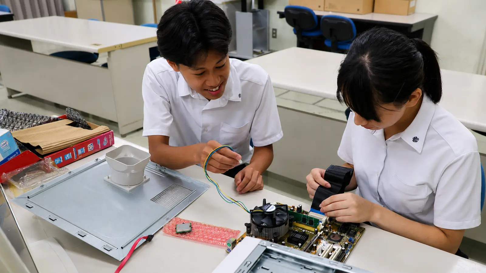 二人の生徒がマザーボードとケーブルを確かめながらパソコンを組み立てる様子
